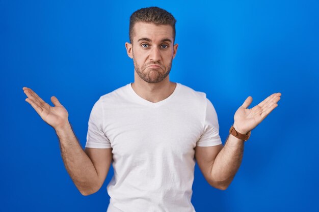 Young caucasian man standing over blue background clueless and confused expression with arms and hands raised doubt concept