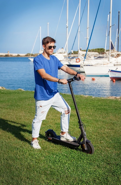 Young Caucasian man stays with an electric scooter on grass near the sea in the marina
