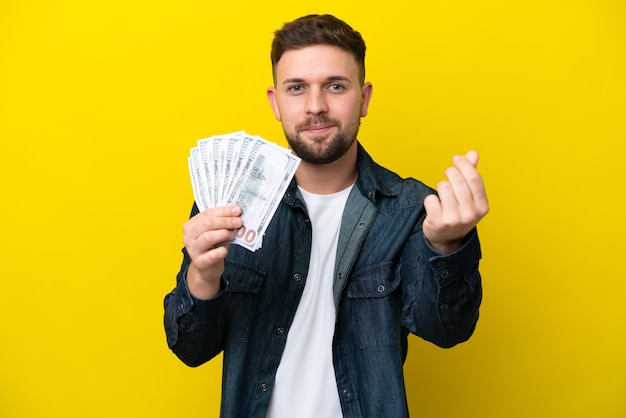 Young caucasian man taking a lot of money isolated on yellow background making money gesture