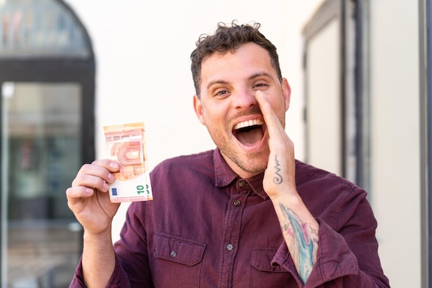 Young caucasian man taking a lot of money at outdoors shouting with mouth wide open