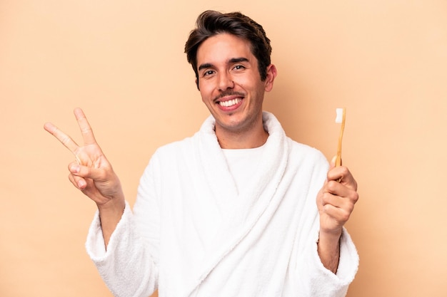 Young caucasian man wearing a bathrobe holding a toothbrush isolated on beige background joyful and carefree showing a peace symbol with fingers