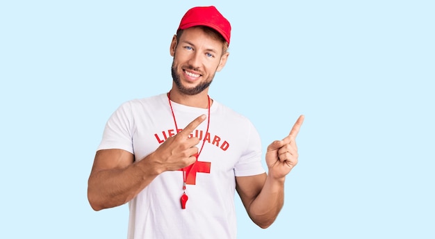 Young caucasian man wearing lifeguard t shirt holding whistle smiling and looking at the camera pointing with two hands and fingers to the side