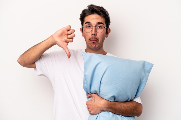 Young caucasian man wearing pajamas holding a pillow isolated on white background showing a dislike gesture, thumbs down. Disagreement concept.