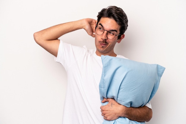 Young caucasian man wearing pajamas holding a pillow isolated on white background touching back of head, thinking and making a choice.