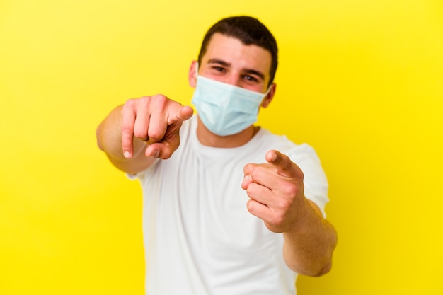 Young caucasian man wearing a protection for coronavirus isolated on yellow background cheerful smiles pointing to front.