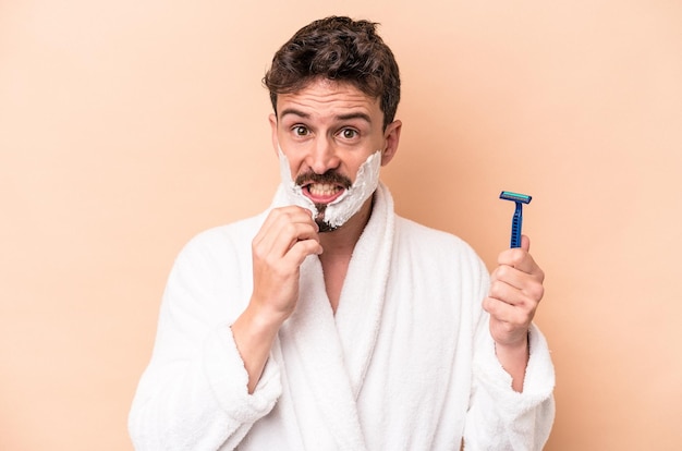 Young caucasian man wearing shaving foam and holding razor blade isolated on beige background biting fingernails, nervous and very anxious.