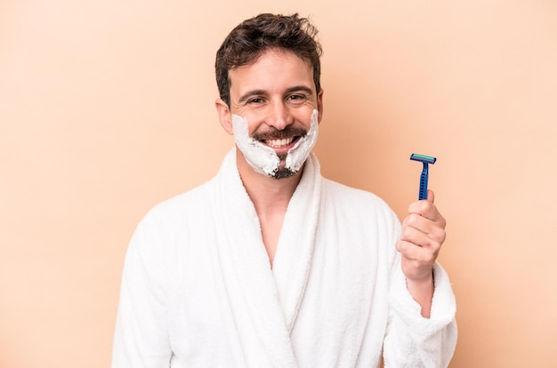 Young caucasian man wearing shaving foam and holding razor blade isolated on beige background happy smiling and cheerful