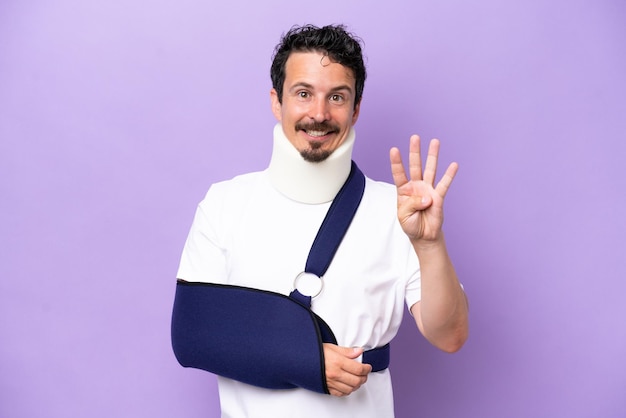 Young caucasian man wearing a sling and neck brace isolated on purple background happy and counting four with fingers