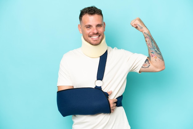 Young caucasian man wearing a sling and neck lace isolated on blue background doing strong gesture