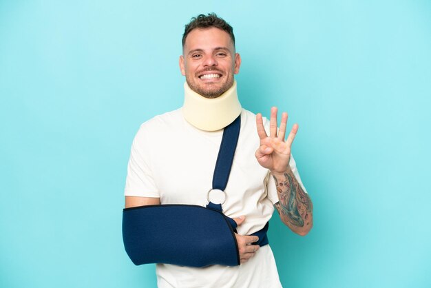 Young caucasian man wearing a sling and neck lace isolated on blue background happy and counting four with fingers