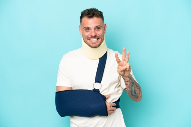 Young caucasian man wearing a sling and neck lace isolated on blue background happy and counting three with fingers