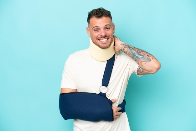 Young caucasian man wearing a sling and neck lace isolated on blue background laughing