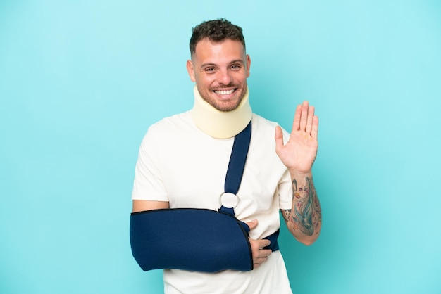 Young caucasian man wearing a sling and neck lace isolated on blue background saluting with hand with happy expression