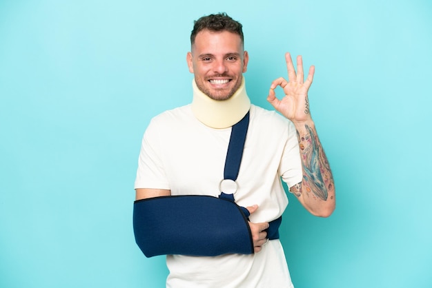 Young caucasian man wearing a sling and neck lace isolated on blue background showing ok sign with fingers