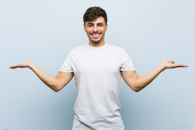 Young caucasian man wearing a white tshirt makes scale with arms, feels happy and confident.