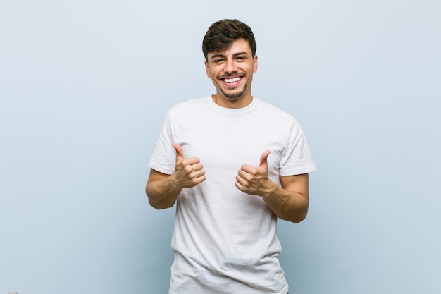 Young caucasian man wearing a white tshirt raising both thumbs up, smiling and confident.