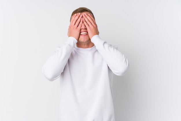 Young caucasian man on white wall covers eyes with hands, smiles broadly waiting for a surprise.