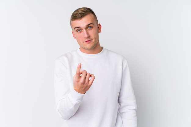 Young caucasian man on white wall pointing with finger at you as if inviting come closer.