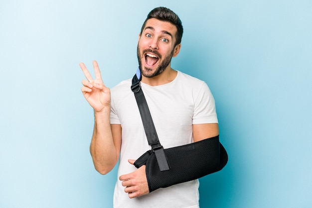 Young caucasian man with broken hand isolated on blue background joyful and carefree showing a peace symbol with fingers