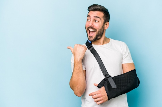 Young caucasian man with broken hand isolated on blue background points with thumb finger away laughing and carefree