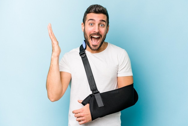 Young caucasian man with broken hand isolated on blue background receiving a pleasant surprise excited and raising hands