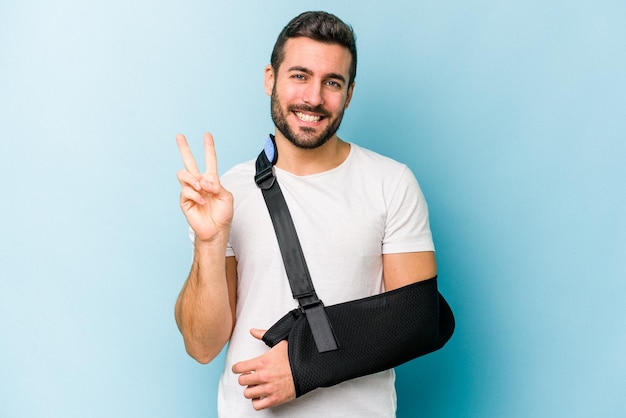 Young caucasian man with broken hand isolated on blue background showing number two with fingers