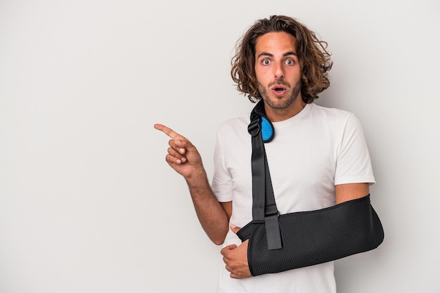 Young caucasian man with broken hand isolated on gray background pointing to the side