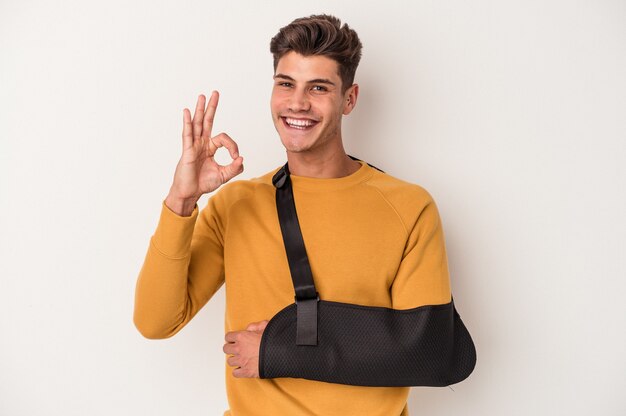 Young caucasian man with broken hand isolated on white background cheerful and confident showing ok gesture.