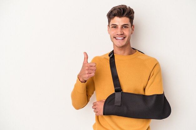 Young caucasian man with broken hand isolated on white background smiling and raising thumb up