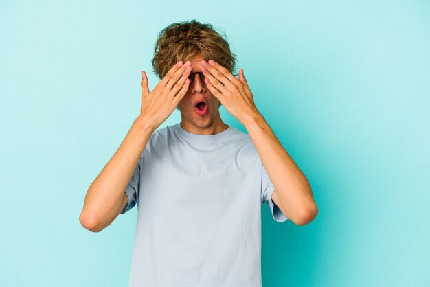 Young caucasian man with make up isolated on blue background  afraid covering eyes with hands.