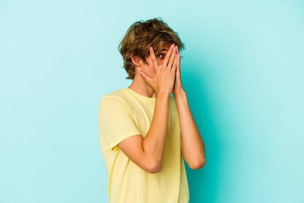 Young caucasian man with make up isolated on blue background  blink through fingers frightened and nervous.