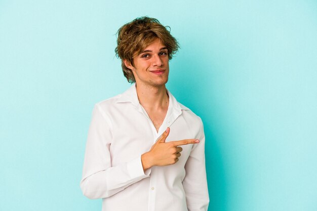 Young caucasian man with make up isolated on blue background  smiling and pointing aside, showing something at blank space.