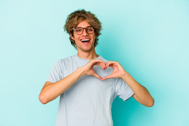 Young caucasian man with make up isolated on blue background  smiling and showing a heart shape with hands.