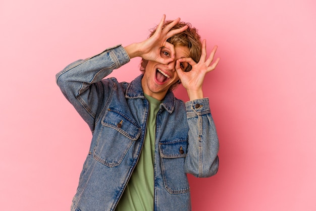 Young caucasian man with make up isolated on pink background showing okay sign over eyes