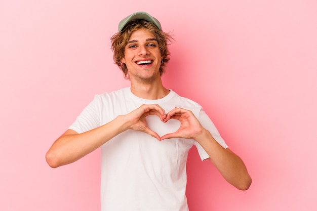 Young caucasian man with make up isolated on pink background smiling and showing a heart shape with hands.