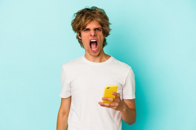 Young caucasian man with makeup holding mobile phone isolated on blue background  screaming very angry and aggressive.