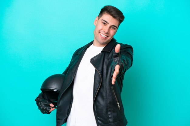 Young caucasian man with a motorcycle helmet isolated on blue background shaking hands for closing a good deal