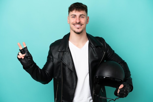 Young caucasian man with a motorcycle helmet isolated on blue background smiling and showing victory sign