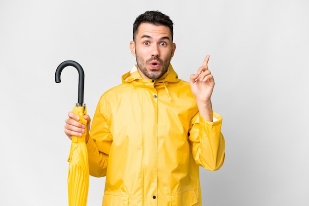 Young caucasian man with rainproof coat and umbrella over isolated white background intending to realizes the solution while lifting a finger up