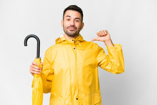 Young caucasian man with rainproof coat and umbrella over isolated white background proud and selfsatisfied