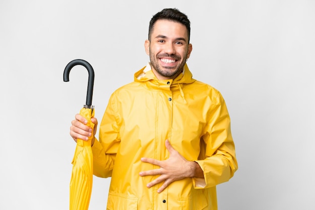 Young caucasian man with rainproof coat and umbrella over isolated white background smiling a lot