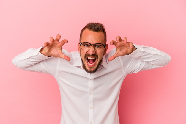 Young caucasian man with tattoos isolated on pink background  showing claws imitating a cat, aggressive gesture.