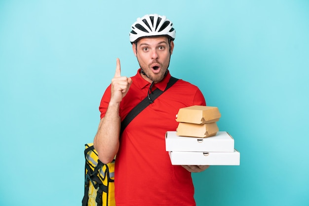 Young caucasian man with thermal backpack and holding fast food isolated on blue background intending to realizes the solution while lifting a finger up