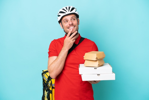 Young caucasian man with thermal backpack and holding fast food isolated on blue background looking up while smiling