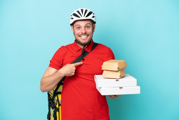 Young caucasian man with thermal backpack and holding fast food isolated on blue background with surprise facial expression