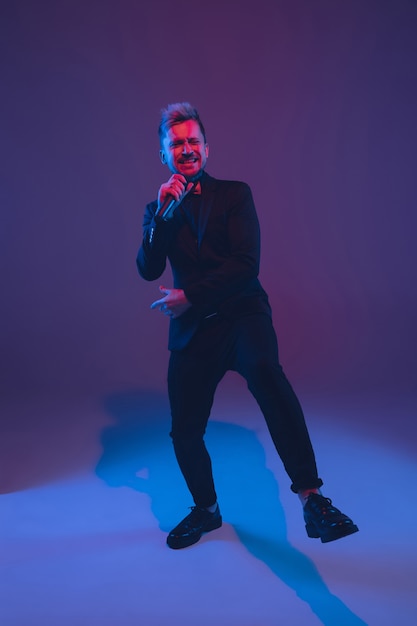 Young caucasian musician, performer singing, dancing in neon light on gradient background