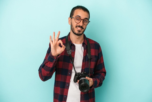 Young caucasian photographer man isolated on blue background cheerful and confident showing ok gesture.