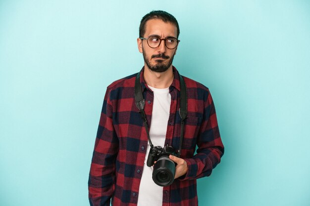 Young caucasian photographer man isolated on blue background confused, feels doubtful and unsure.