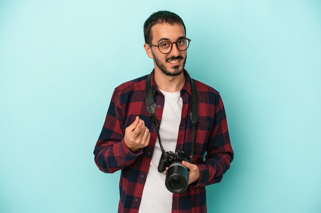 Young caucasian photographer man isolated on blue background pointing with finger at you as if inviting come closer.