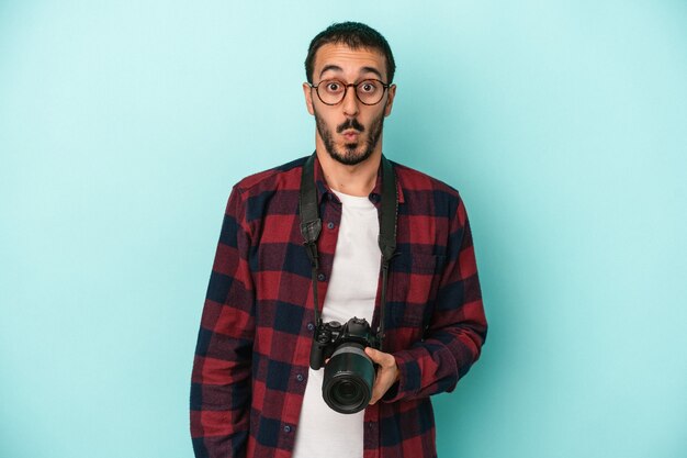 Young caucasian photographer man isolated on blue background shrugs shoulders and open eyes confused.